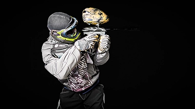 Nahaufnahme eines Paintball-Athleten, der seinen Markierer hält und bereit zum Schießen ist. Sein Gesicht ist durch die Maske verdeckt, aber seine Körperhaltung strahlt Konzentration und Entschlossenheit aus. Das Bild ist vor einem komplett schwarzen Hintergrund aufgenommen.